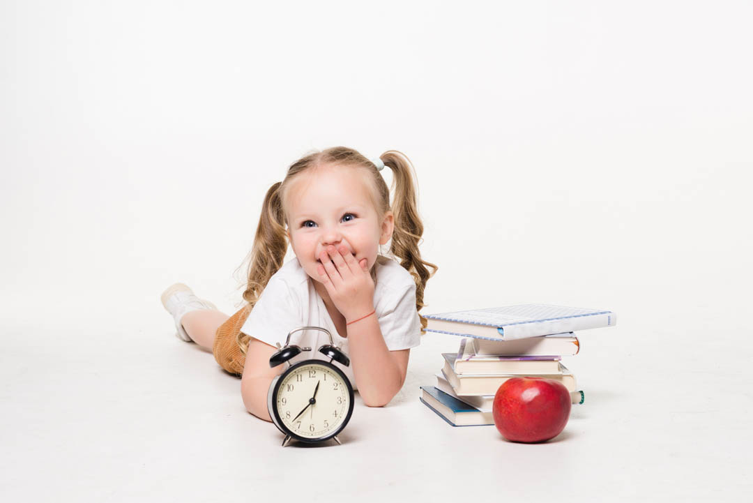 Education and school concept. Smiling little student girl with book clock and apple lying on the floor