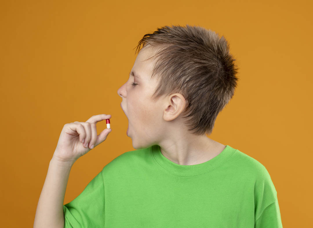 ill little boy in green t-shirt feeling unwell taking a pill standing over orange background